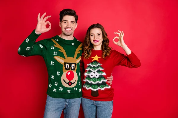 Portret van vrolijke twee mensen student met brunette golvende haar knuffel Embrace Toon OK teken verkoop slijtage grappige herten kerstboom Pullover denim jeans geïsoleerd over rode kleur achtergrond — Stockfoto