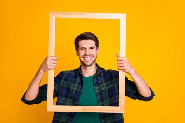 Foto de tipo divertido sosteniendo marco de madera mirando a través de ella posando ropa casual camisa a cuadros aislado color amarillo fondo — Foto de Stock