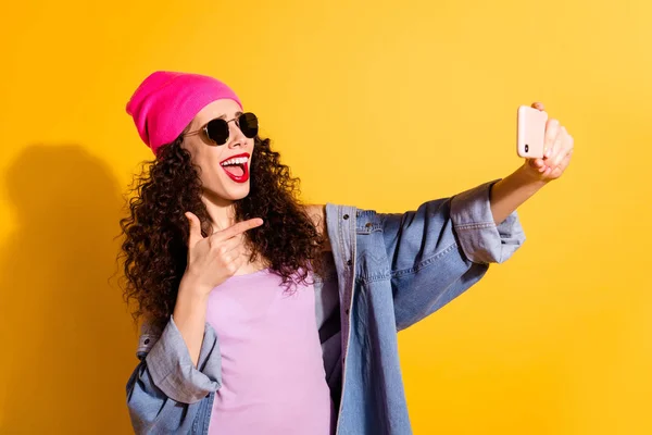 Foto de senhora ondulada elegante segurando telefone fazendo selfies dirigindo dedo para câmera usar roupas casuais isolado cor amarela fundo — Fotografia de Stock