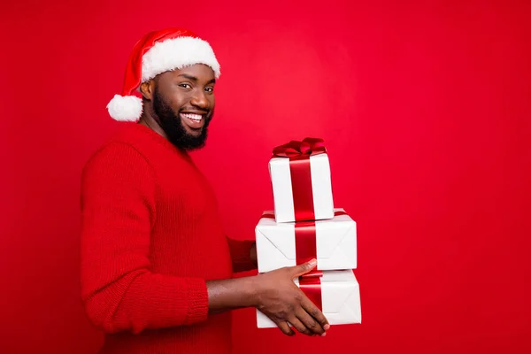 Retrato de homem alegre com muitos pacotes carregando na noite de Natal vestindo chapéu de santa claus boné usando suéter isolado sobre fundo vermelho — Fotografia de Stock