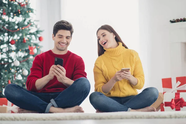 Foto de longitud completa de dos personas alegres utilizan el hombre del teléfono celular muestran x-mas descuentos estacionales felicitaciones con la Navidad sentarse piso de tonelada en casa con ambiente de decoración de año nuevo — Foto de Stock