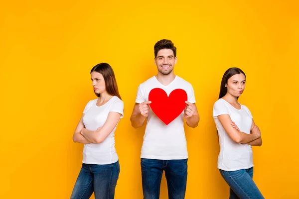 Retrato de un chico alegre alegre lindo atractivo agradable sosteniendo en las manos gran corazón decidiendo entre dos novias ofendidas aisladas sobre brillante brillante brillo fondo amarillo — Foto de Stock