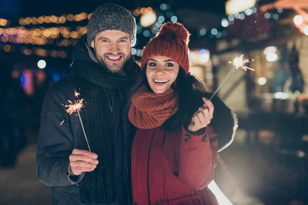 Foto de casal engraçado na celebração x-mas no parque segurando faíscas mágicas animado para atender a meia-noite do ano novo usando casacos quentes de malha bonés e lenços ao ar livre — Fotografia de Stock