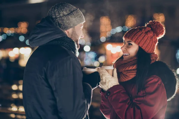 Foto von zwei Personen mit heißem Teegetränk in der Hand, die den Weihnachtsabend im dekorierten Park verbringen, während sie draußen warme Mäntel tragen — Stockfoto