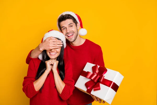 クリスマスキャップの魅力的な男の肖像画は、彼の素敵な恋人の目を隠し、彼女のパッケージは黄色の背景の上に孤立した赤いセーターを着用します — ストック写真
