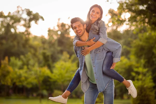 Portret Wesoła małżonków z Brunet włosów piggyback noszenia jeansów w parku na zewnątrz — Zdjęcie stockowe