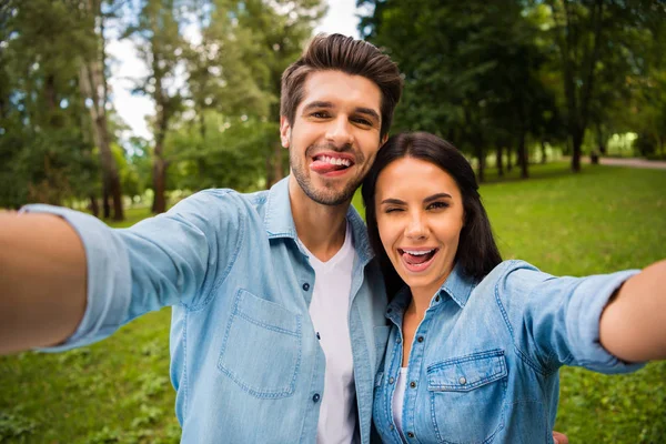 Tutup foto pasangan yang kekanak-kanakan dengan rambut cokelat membuat foto yang menunjukkan lidah mereka berkedip sambil mengenakan kaus jeans denim di luar ruangan — Stok Foto