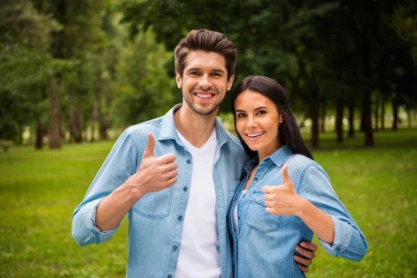 Retrato de casal encantador com cabelo morena mostrando polegar para cima sorrindo vestindo jeans camisas jeans jaquetas fora na floresta — Fotografia de Stock