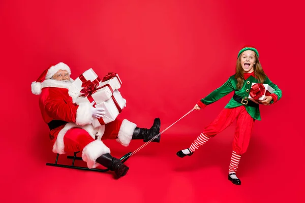 Photo pleine grandeur de santa claus drôle avec lunettes lunettes lunettes et elfe mignon en chapeau vert coiffure tenant traîneaux portent sac avec des cadeaux isolés sur fond rouge — Photo