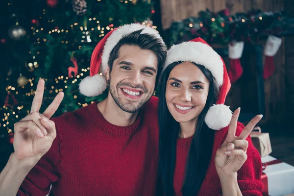 キャップハットを持つ2人の結婚したロマンスロマンチックな人々のクローズアップ写真は、Vサインは、屋内でノエルの装飾イルミネーションの新しい年のライトの完全な家でクリスマス休暇X-masの休日を楽しみます — ストック写真
