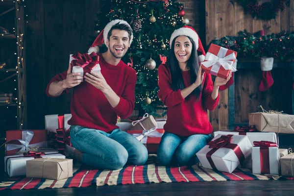 Foto de corpo inteiro de dois cônjuges espantados tem chapéu de boné de Papai Noel sentar no cobertor xadrez mostrar presentes x-mas para a noite de Natal em casa com luzes de decoração noel dentro de casa — Fotografia de Stock