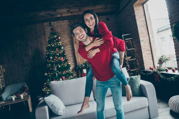 Portrait of romantic two youth hug piggyback on christmas x-mas party event wearing red jumper denim jeans in house with decoration illumination lights indoors near couch