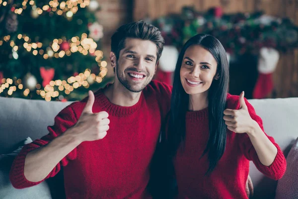 Retrato da linda morena de cabelos dois cônjuges mostrando polegar para cima sentado no divã em interior decorativo com iluminação x-mas ano novo em casa — Fotografia de Stock