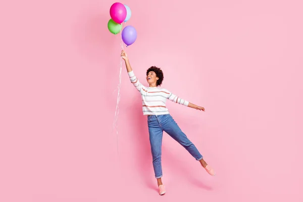 Foto de larga duración de la bella mujer ondulada de piel oscura sosteniendo globos de aire de colores va a volar con el viento soplando desgaste jersey de rayas aislado pastel de color rosa de fondo —  Fotos de Stock