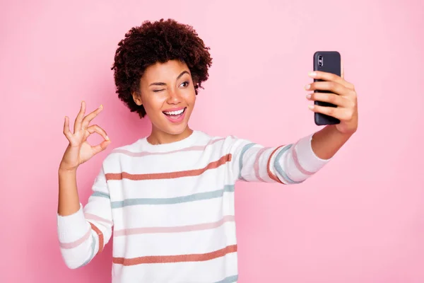 Photo de jolie dame de peau sombre tenant téléphone faisant selfies montrant okey symbole clin d'oeil usure rayé pull isolé couleur rose pastel fond — Photo