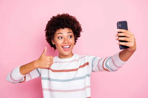 Foto de la señora de piel oscura bastante excitada sosteniendo el teléfono haciendo selfies levantando el pulgar hacia arriba usar suéter de rayas blancas aislado pastel color rosa fondo —  Fotos de Stock