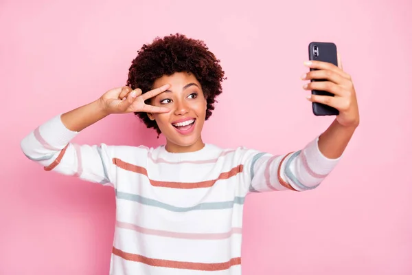 Foto de positiva alegre linda encantadora fascinante hermosa atractiva novia de piel oscura que le muestra v-signo de tomar selfie con suéter de rayas blancas aisladas sobre el color pastel rosa —  Fotos de Stock