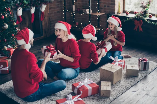 Фотография всей семьи папа мама сестра брат обмениваясь рождественскими подарками сидя уютный ковёр на полу рядом с украшенными гирляндами огни новогодней елки в помещении носить Санта шапки красные джамперы — стоковое фото