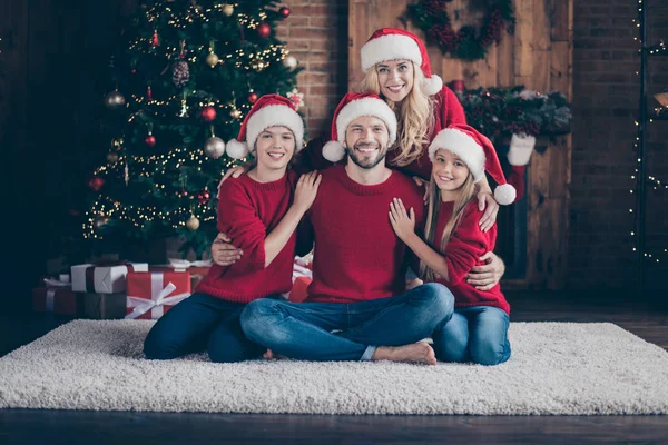 Foto de grandes quatro membros família pai mãe irmã irmão passar x-mas véspera juntos sentado chão perto de guirlanda árvore dentro de casa usar bonés de Papai Noel e camisolas vermelhas — Fotografia de Stock