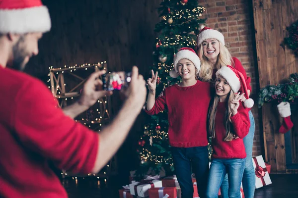 Photo de papa faisant la photo de frère soeur et maman montrant v-signe près décoré nouvel an arbre intérieur x-mas esprits porter santa casquettes et pulls rouges — Photo