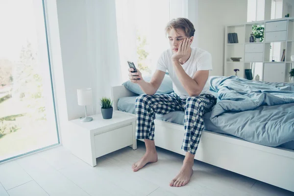 Full length photo of tired unhappy upset blonde haired man have night work try not sleep use his cellphone follow social network accounts blogs sit on bed in house indoors