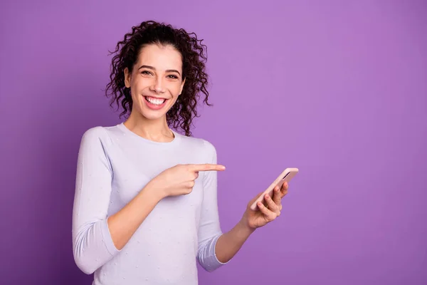 Foto de linda mujer encantadora alegre linda creando copyspace apuntando a su teléfono espacio vacío sostenido con las manos usando suéter aislado sobre fondo de color pastel violeta — Foto de Stock