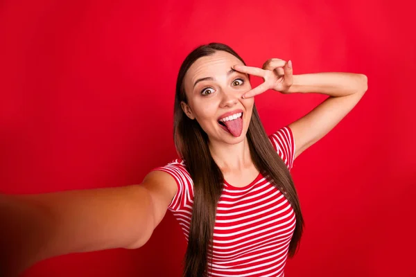 Автопортрет веселої милої милої чарівної досить модної дивовижної дівчини, яка показує вам язик, що посміхається в красивій смугастій футболці, одночасно ізольовані на червоному тлі — стокове фото