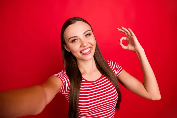 Автопортрет чарівної милої красивої привабливої дівчини, що показує вам знак ок під час носіння смугастої футболки ізольовано червоним тлом — стокове фото