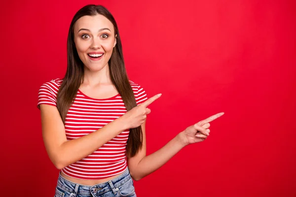 Photo of charmiung nice cute astonished girl wearing jeans denim adverting you something cool by pointing at it while isolated with red background