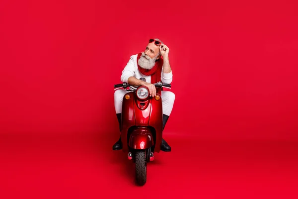 Фотографія повної довжини вдумливої літньої людини, яка торкається окулярів, сидить на велосипеді Подумайте про те, щоб носити білі штани-перемички ізольовані на червоному тлі — стокове фото