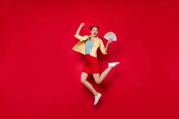 Πηδώντας ψηλά περιχαρής κυρία κρατώντας ανεμιστήρα του δολάρια χέρια φορούν casual στολή απομονωμένο κόκκινο φόντο — Φωτογραφία Αρχείου