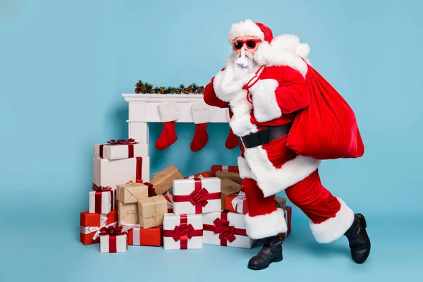 Ganzkörperansicht seines schönen fetten geheimnisvollen bärtigen Weihnachtsmannes, der große Säckelstapel mit Einkäufen trägt, zeigt das Zeichen shh isoliert über blauem, pastellfarbenem Hintergrund — Stockfoto