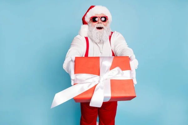 Modern gözlüklü komik, aşırı kilolu Noel Baba portresi Noel arifesinde büyük bir hediye kutusu beyaz kurdelesi verir Noel arifesinde kırmızı pantolon askısı giyer mavi arka plan üzerine izole edilmiş eldivenler giyer. — Stok fotoğraf