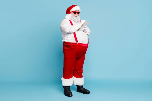 Pleine longueur photo de drôles funky graisse santa claus dans les lunettes hipster avec gros ventre écrire liste de souhaits plan dans le livre note utilisation stylo porter bretelles pantalons rouges bottes isolées sur fond de couleur bleue — Photo