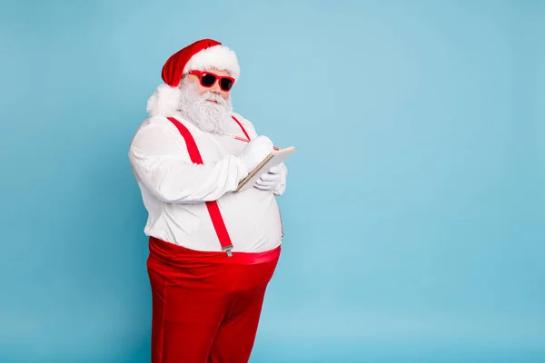Girato foto di divertente funky grasso Babbo Natale in occhiali moderni con pancia grande addome scrivere con la matita nel libro nota fare lista dei desideri indossare bretelle rosse isolate su sfondo di colore blu — Foto Stock