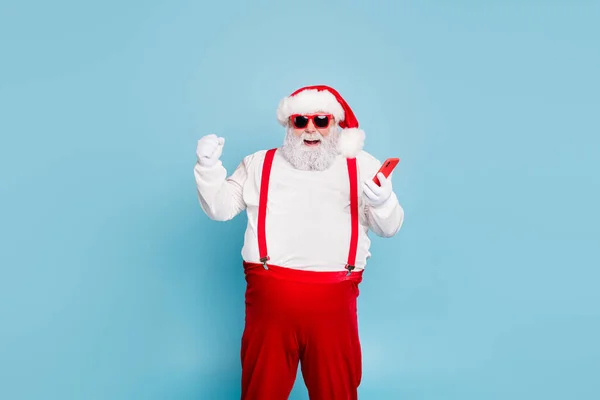 Portré vicces funky őrült karácsony apa nagy hasa használata mobiltelefon emel öklöt sikoly igen győzelem x-mas téli vásárlás lottó viselni piros nadrágtartó elszigetelt kék színű háttér — Stock Fotó