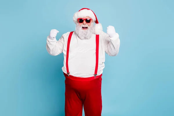 Портрет смешной фанки толстый Санта-Клаус с большим животом поднять кулаки кричать да удачи праздновать новогоднюю рождественскую вечеринку носить подтяжки современные красные очки изолированы на синем фоне цвета — стоковое фото