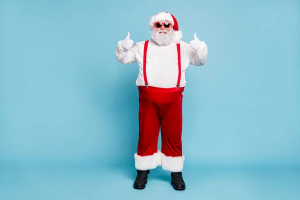 Foto de corpo inteiro de engraçado funky gordura Papai Noel com barriga grande abdômen em óculos mostrar polegar para cima recomendar inverno temporada compras anúncios usar macacão vermelho botas calças isoladas sobre fundo de cor azul — Fotografia de Stock