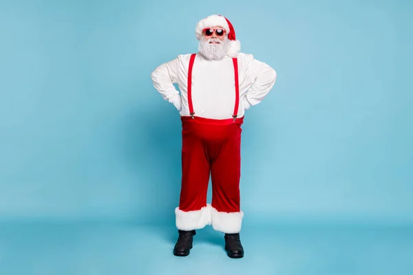 Foto de longitud completa de divertidos funky grasa con sobrepeso de pelo gris santa claus con sombrero gafas modernas disfrutar de Navidad tiempo usar monos guapos pantalones rojos botas aisladas sobre fondo de color azul —  Fotos de Stock