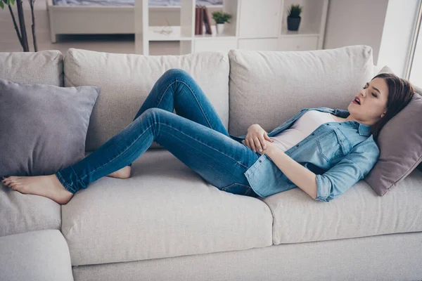 Full length photo of upset girl lying on sofa touch her tummy suffer from pain in belly have terrible pms wear denim jeans outfit in room apartment — ストック写真