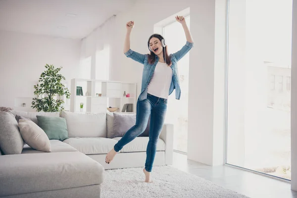 Полнометражное фото удивительной леди, слушающей любимую мелодию в современных ушанках, танцующей в светлой комнате в джинсовой одежде, квартира в помещении — стоковое фото