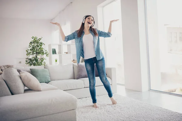 Foto de larga duración de la increíble dama escuchando melodía favorita en las orejeras modernas levantando las manos bailando encantado en la sala de luz con pantalones vaqueros ropa apartamento interior — Foto de Stock