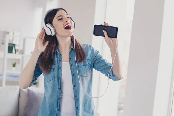 Foto av söt dam med telefon i händerna öronlappar på öronen sjunger högt hemtrevligt humör står nära soffan bär jeans kläder lägenhet inomhus — Stockfoto