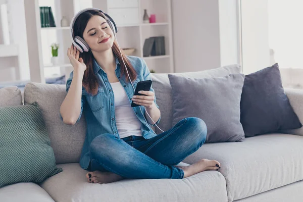 Photo of pretty dreaming lady with telephone in hands earflaps on ears listening new radio fm sitting comfy sofa wearing jeans clothes apartment indoors