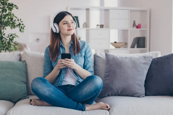 Foto av söt drömmande dam med telefon i händerna slå på cool modern sång i öronlappar sitter bekväm soffa bär jeans kläder lägenhet inomhus — Stockfoto