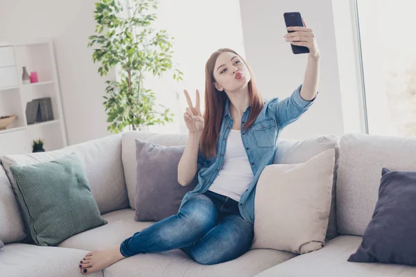 手の中に浮気女性の電話の写真自己紹介vサインシンボル送信空気キス座って快適な上のソファ着てジーンズ服アパート室内 — ストック写真