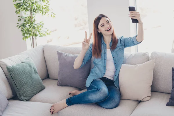 Photo de jolie dame avec téléphone dans les mains faisant selfies montrant le symbole de v-signe assis confortablement sur le canapé portant des vêtements de jeans appartement à l'intérieur — Photo