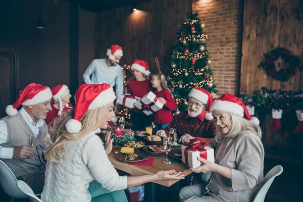 Genç insanların mutlu noel aile toplantısı küçük erkek çocuklar olgun emekliler yeni yıl ziyafeti çeken kadınla masaya oturur annesinin Noel Baba şapkası taktığı hediye kutusunu verir evde Noel Baba şapkası partisinin tadını çıkar. — Stok fotoğraf