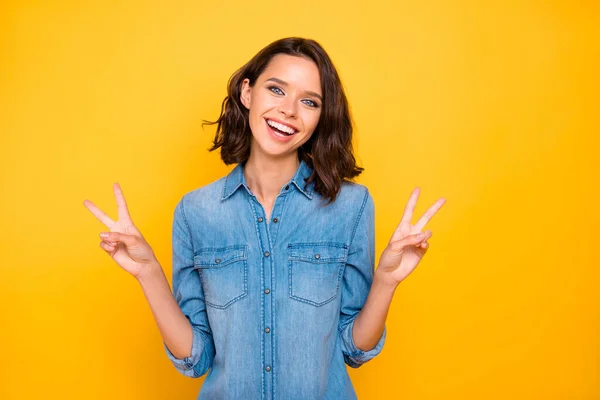 Retrato de positiva alegre encantadora chica disfrutar del tiempo libre hacer v-signos de desgaste jeans camisa aislada sobre fondo de color amarillo brillante —  Fotos de Stock