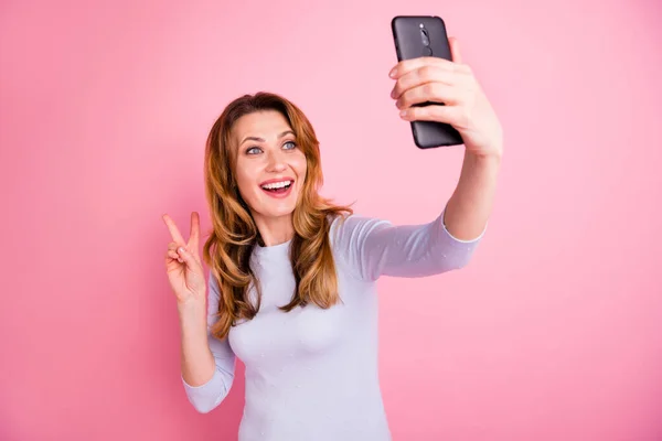 休日の肯定的な陽気な女性の肖像画は、彼女の携帯電話で自画自身を取りますVサインは、コンテンツがピンクの色の背景に隔離された白いジャンパーを着用感じます — ストック写真